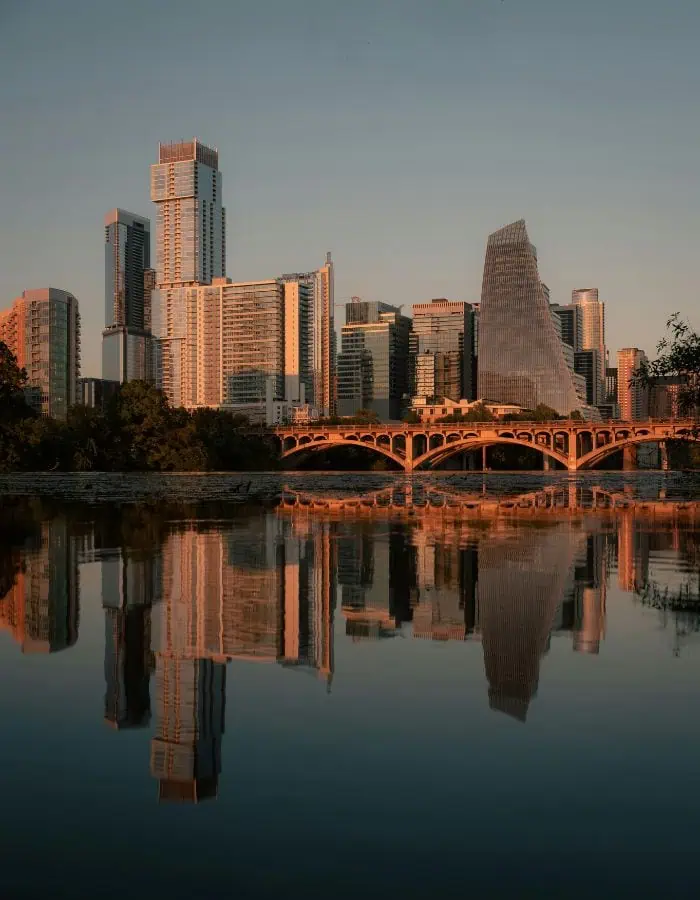 Modern skyscrapers of downtown Austin, Texas glow in the warm light of sunset, with the arched Ann W. Richards Congress Avenue Bridge spanning across the calm Colorado River. The buildings and bridge are mirrored perfectly in the still water, creating a striking cityscape reflection.