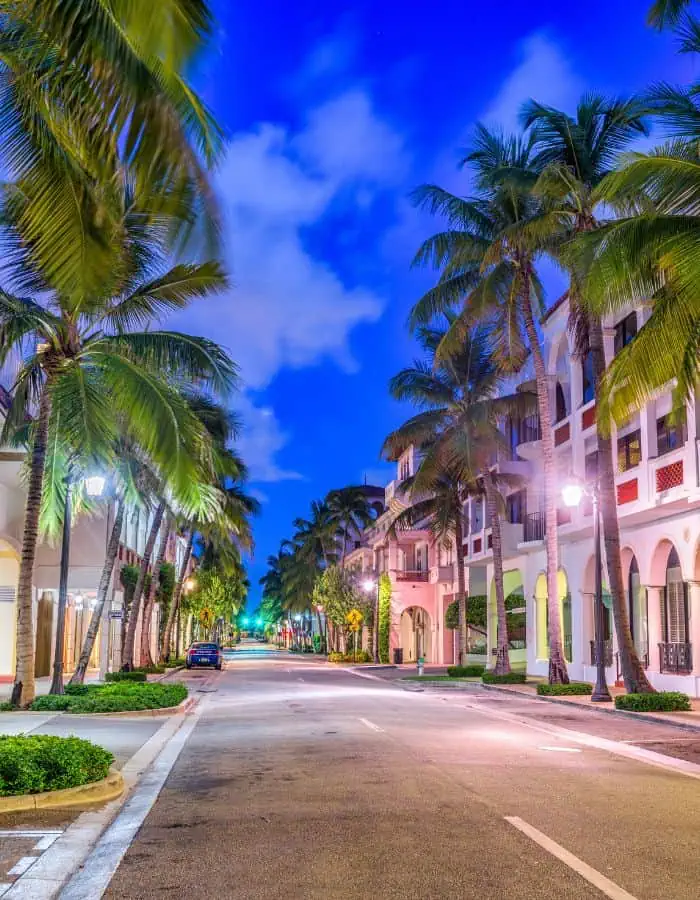 A quiet, palm-lined street in Palm Beach, Florida at dusk, with pastel-colored Mediterranean-style buildings on either side. The sky glows a vibrant blue with scattered clouds, and streetlights cast a soft glow on the empty road.
