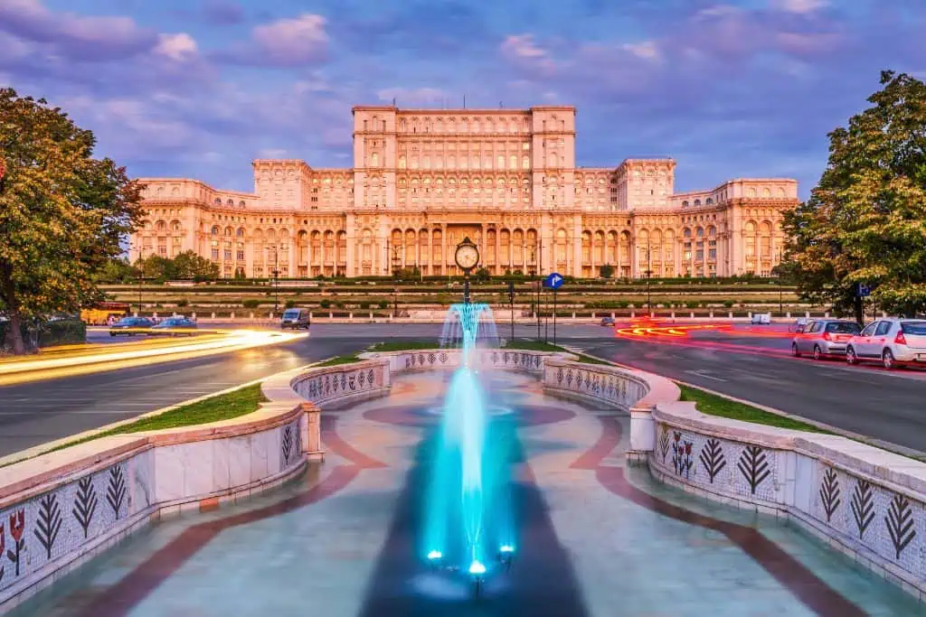 Grand view of the Palace of the Parliament in Bucharest, Romania, illuminated in soft golden light at sunset. A vibrant blue fountain and mosaic-tiled water feature lead toward the imposing neoclassical building, with car light trails curving along the road on both sides.