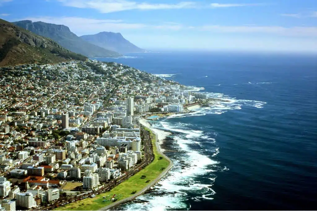 Aerial view of Cape Town, South Africa, with the city sprawling along the rugged coastline where waves crash against the shore. Table Mountain and the Twelve Apostles rise in the background, while a mix of high-rise buildings and suburban homes line the scenic waterfront.