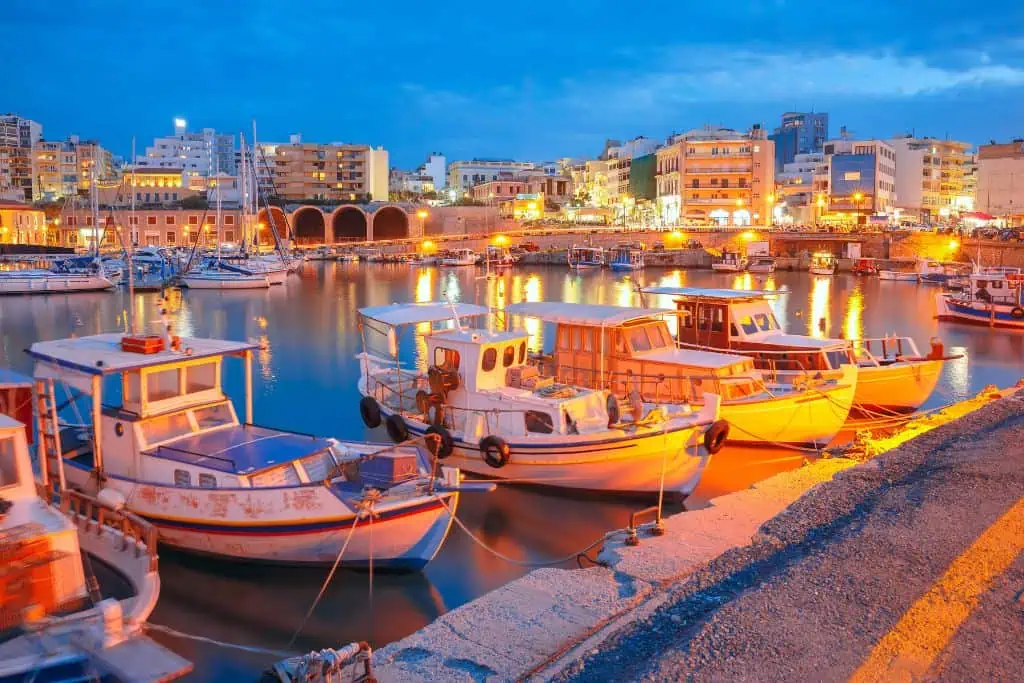 Colorful fishing boats float peacefully in the softly lit harbor of Heraklion, Crete, Greece, as evening falls over the city. Warm golden lights from waterfront buildings reflect on the calm water, with a backdrop of modern apartments and historic arched shipyards under a dusky blue sky.