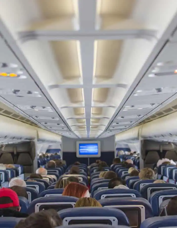 View from the back of a commercial airplane cabin showing passengers seated, overhead compartments closed, and an in-flight screen displaying information at the front of the cabin.