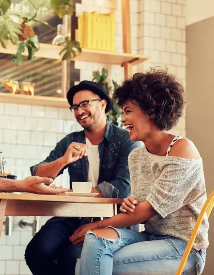 Friends laugh and enjoy a lively conversation at a cozy café table, with coffee cups in front of them and another person gesturing from out of frame. The setting is warm and stylish, with tiled walls, plants, and bright natural light adding to the cheerful atmosphere.