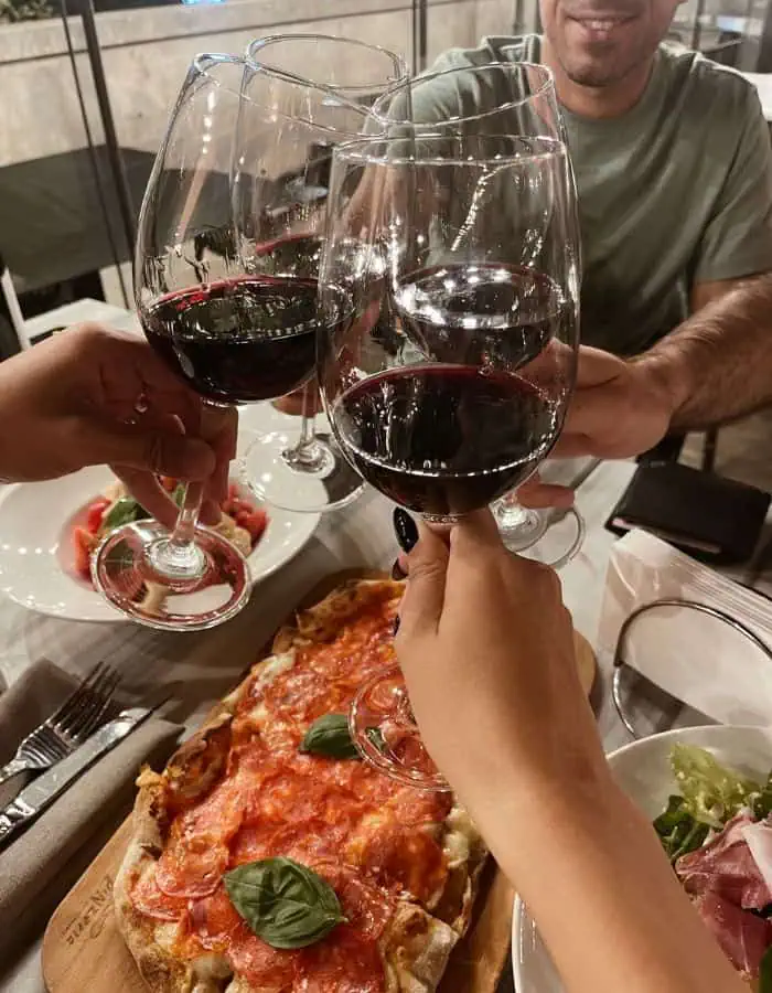 Four people clink glasses of red wine over a shared dinner table, smiling and enjoying a cozy meal. A rectangular pepperoni pizza garnished with fresh basil sits at the center, surrounded by plates of salad and sliced tomatoes with mozzarella.