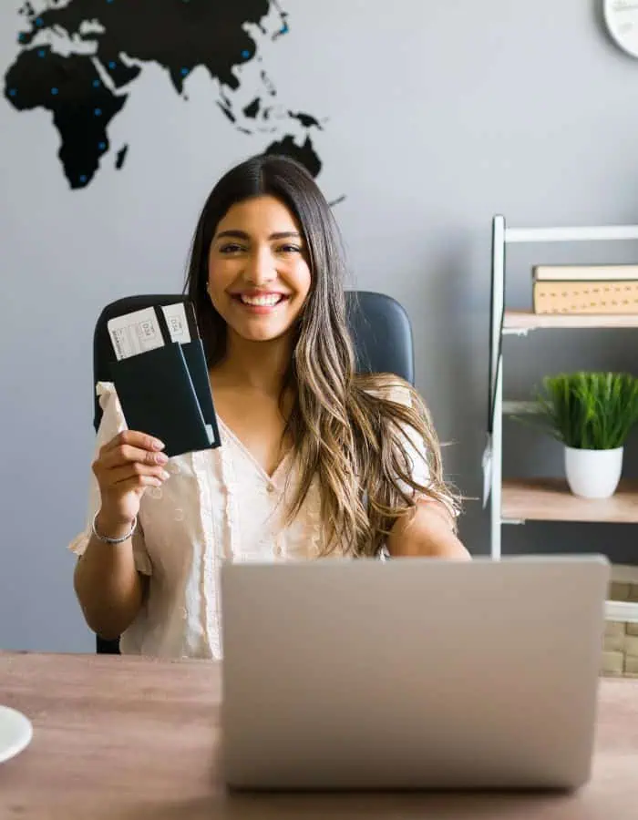 Smiling woman sitting at a desk with a laptop, holding up a passport and airline tickets, with a world map on the wall behind her, suggesting she's ready to travel.