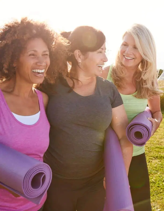 Three women smile and chat as they walk outdoors in the sunlight, each carrying a rolled-up purple yoga mat. Dressed in casual workout clothes, they appear relaxed and happy, suggesting a post-yoga glow or a friendly meetup for a fitness session.