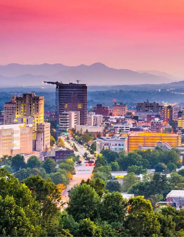A colorful sunset casts a warm glow over downtown Asheville, North Carolina, with lush green trees in the foreground and the Blue Ridge Mountains layered in the background. The city’s mix of modern and historic buildings lines a central road that leads toward the horizon.