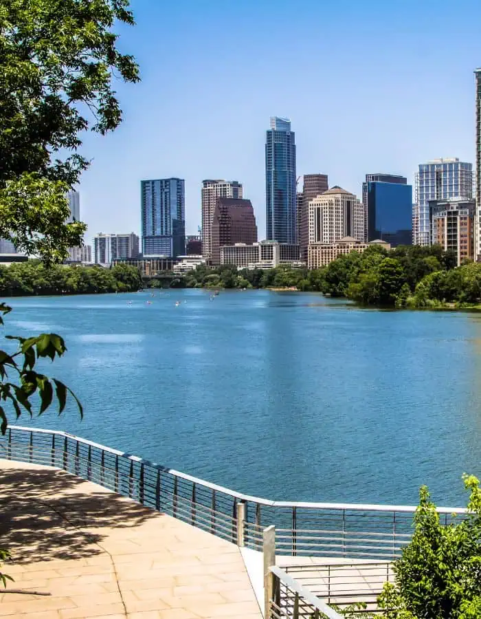 A view of downtown Austin, Texas, showcases modern skyscrapers rising above the calm blue waters of Lady Bird Lake. Framed by green trees and a riverside walkway in the foreground, the scene reflects a mix of urban life and natural beauty on a sunny day.