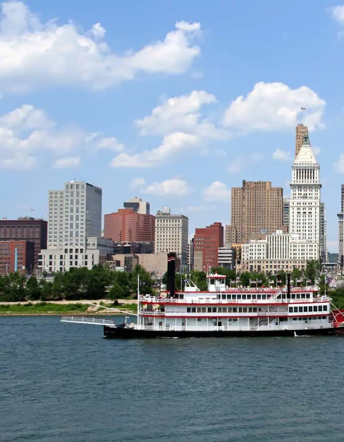 A classic riverboat with red and white trim floats on the Ohio River in front of the Cincinnati skyline, featuring a mix of modern and historic buildings under a partly cloudy blue sky. The scene captures the city's riverfront charm and architectural variety.