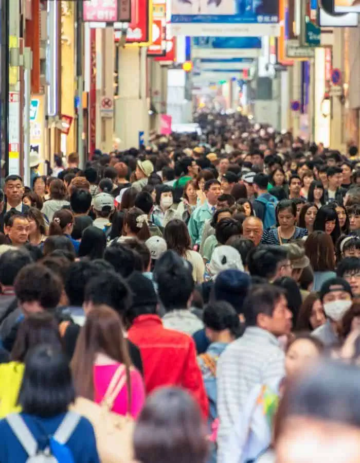 A densely packed crowd of people walks through a busy, covered shopping street lined with colorful signs and advertisements in an urban setting. The image captures the hustle and energy of city life, with some individuals wearing masks and others engaged in conversation.