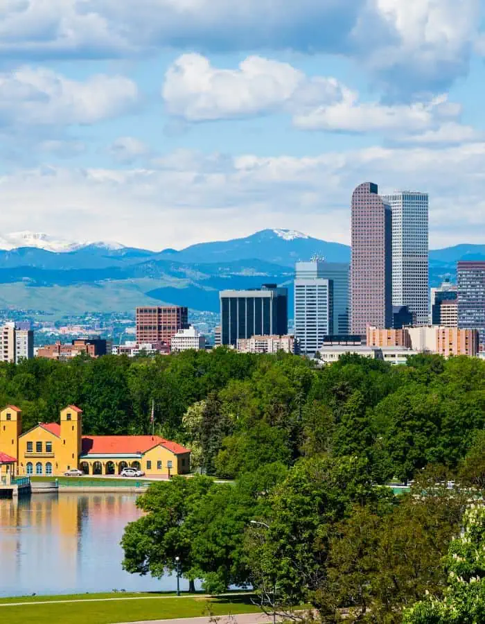 The Denver, Colorado skyline rises behind a lush expanse of trees and a small lake, with the iconic yellow pavilion at City Park in the foreground. Snow-capped Rocky Mountains create a dramatic backdrop under a partly cloudy sky, highlighting the city’s unique mix of urban and natural beauty.