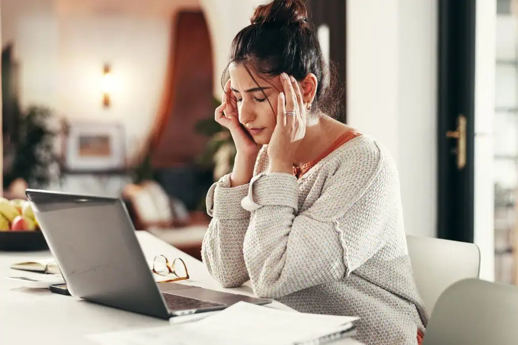 Woman sits at a table with her laptop, holding her head in her hands as if suffering from digital nomad burnout. Glasses and paperwork lie in front of her in a softly lit home setting.