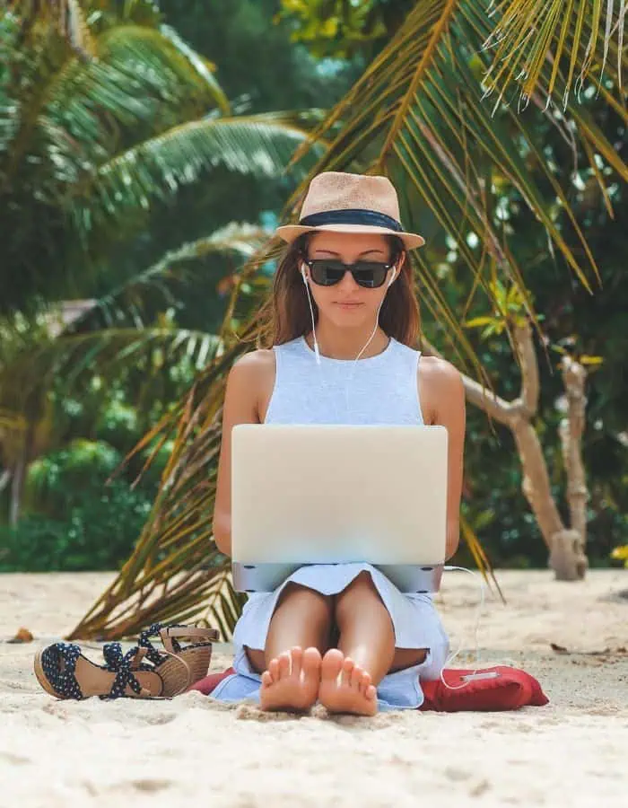 A woman sits barefoot on a sandy beach under palm trees, wearing a sunhat and sunglasses while working on a laptop with earphones in. Her wedge sandals are placed beside her, emphasizing a relaxed yet focused remote work setting in a tropical location.