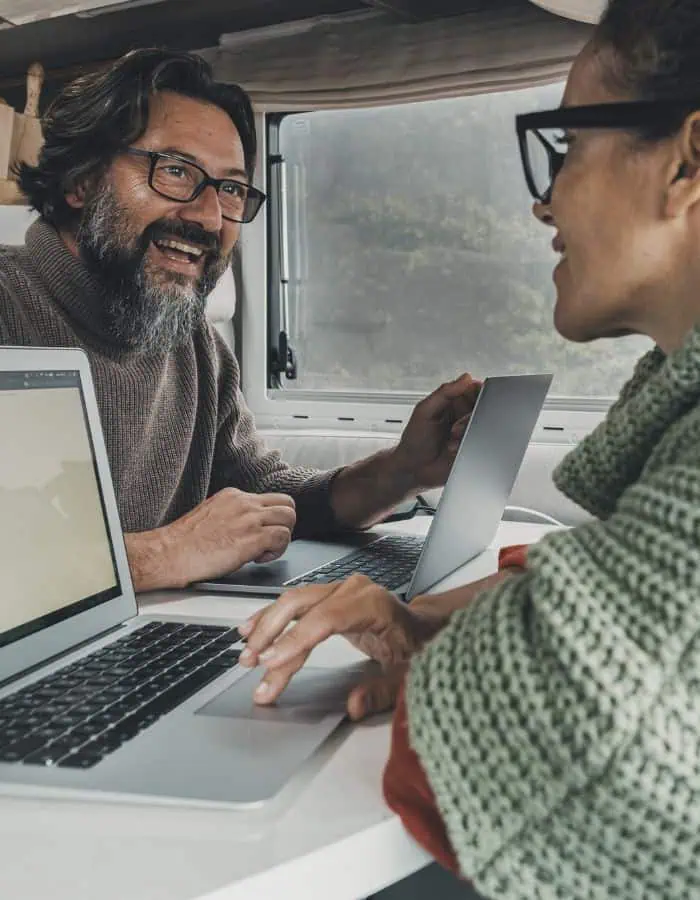 A man and woman sit across from each other inside a camper van, both working on laptops and smiling in conversation. Natural light filters through the window, creating a cozy and collaborative remote work atmosphere on the road.