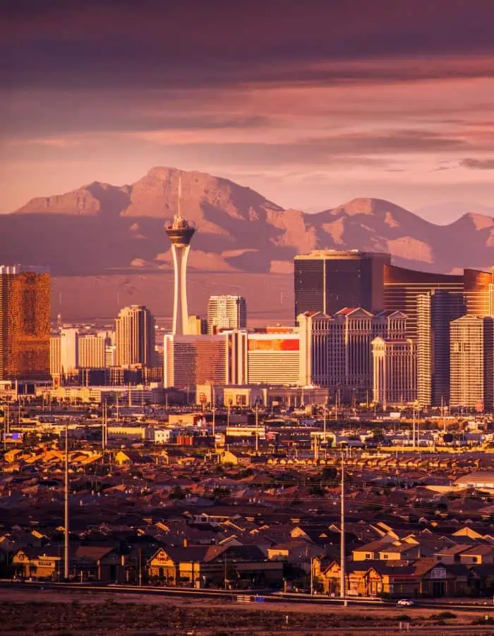 The Las Vegas skyline glows under a dramatic sunset, with the Stratosphere Tower standing tall among modern hotels and casinos. In the foreground, a dense grid of suburban homes stretches toward the glittering Strip, backed by rugged desert mountains.