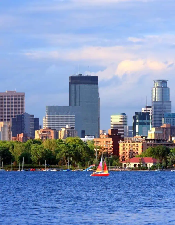 A colorful sailboat glides across a calm lake with the Minneapolis skyline rising in the background under a partly cloudy sky. Lush green trees and low-rise buildings line the waterfront, contrasting with the modern high-rises beyond.