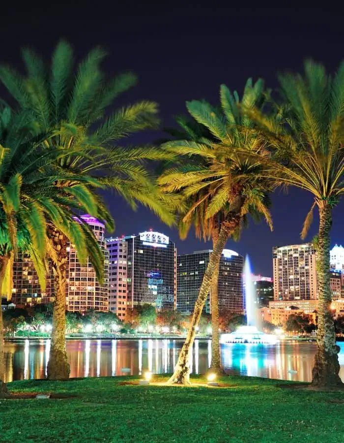 Tall palm trees frame a glowing cityscape at night in downtown Orlando, Florida, with colorful building lights reflected in the still waters of Lake Eola. A lit fountain adds a striking focal point in the lake, enhancing the vibrant and tropical urban setting.
