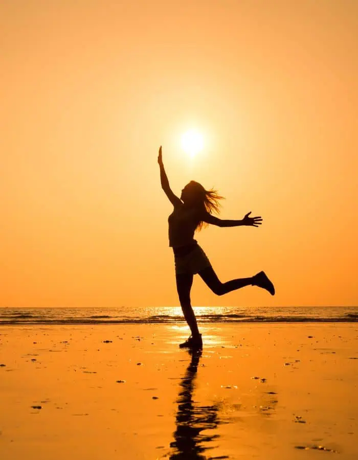 Silhouette of a woman joyfully leaping on a beach at sunset, with her arms raised and the sun directly above her hand. The golden sky and reflective wet sand create a warm, glowing backdrop that emphasizes the carefree mood.