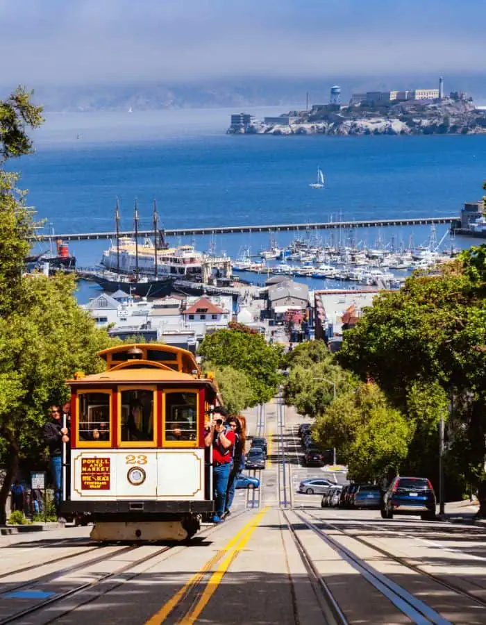 A classic San Francisco cable car travels downhill toward the waterfront, with Alcatraz Island visible in the distance across the bay. Lush trees line the steep street, and the marina filled with sailboats adds to the city's iconic coastal charm.