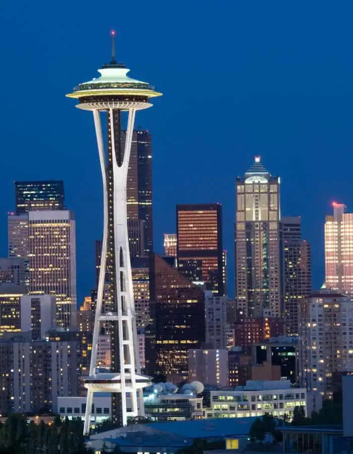 The iconic Space Needle stands illuminated against the Seattle skyline at night, surrounded by a cluster of high-rise buildings glowing with city lights. The deep blue sky enhances the striking contrast and modern architecture of this Pacific Northwest city.
