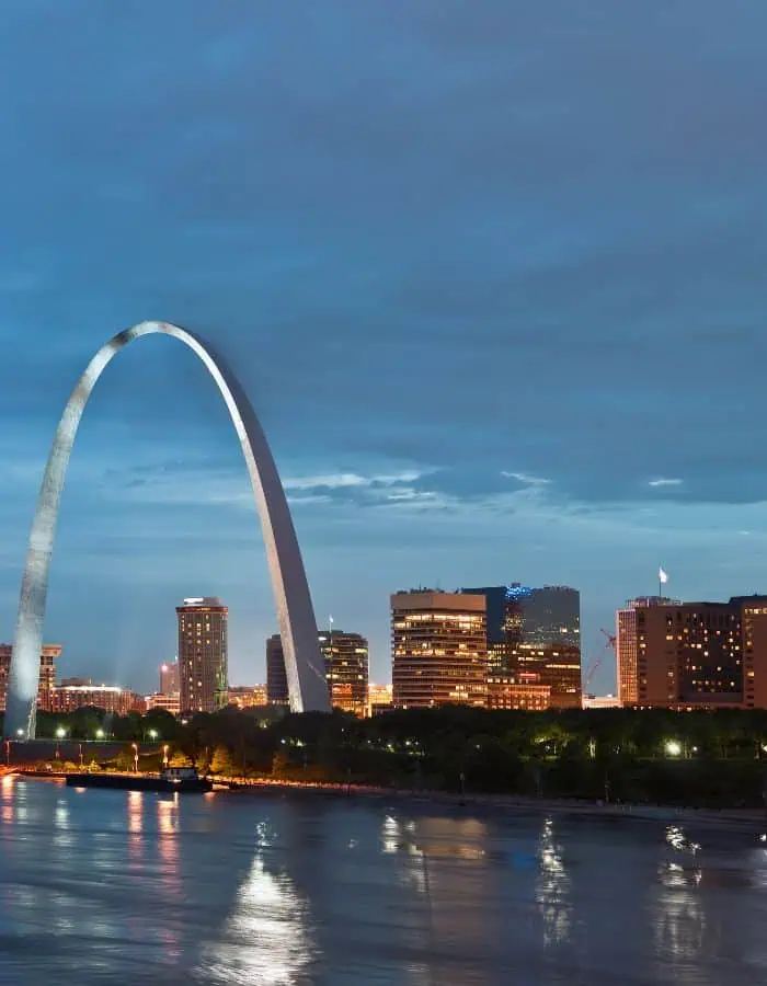 The Gateway Arch rises gracefully over the St. Louis skyline at dusk, illuminated against a backdrop of modern buildings and a cloudy blue sky. Reflections from city lights shimmer across the surface of the Mississippi River in the foreground.