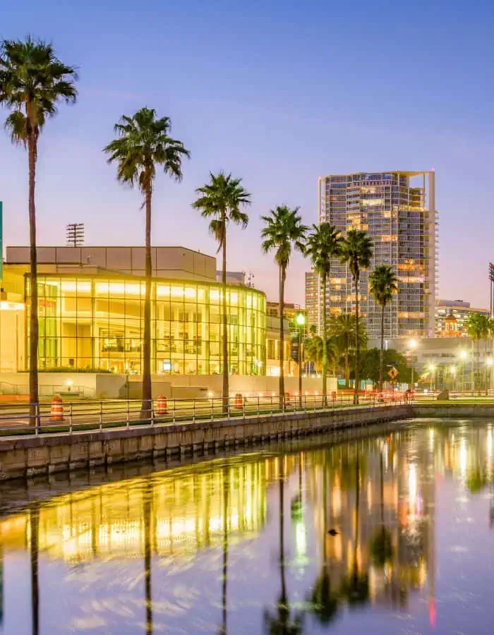 A twilight view of downtown St. Petersburg, Florida, features a modern, glass-walled building glowing with interior lights beside a calm waterfront. Tall palm trees and high-rise apartments are reflected in the water, adding to the warm, tropical ambiance of the cityscape.