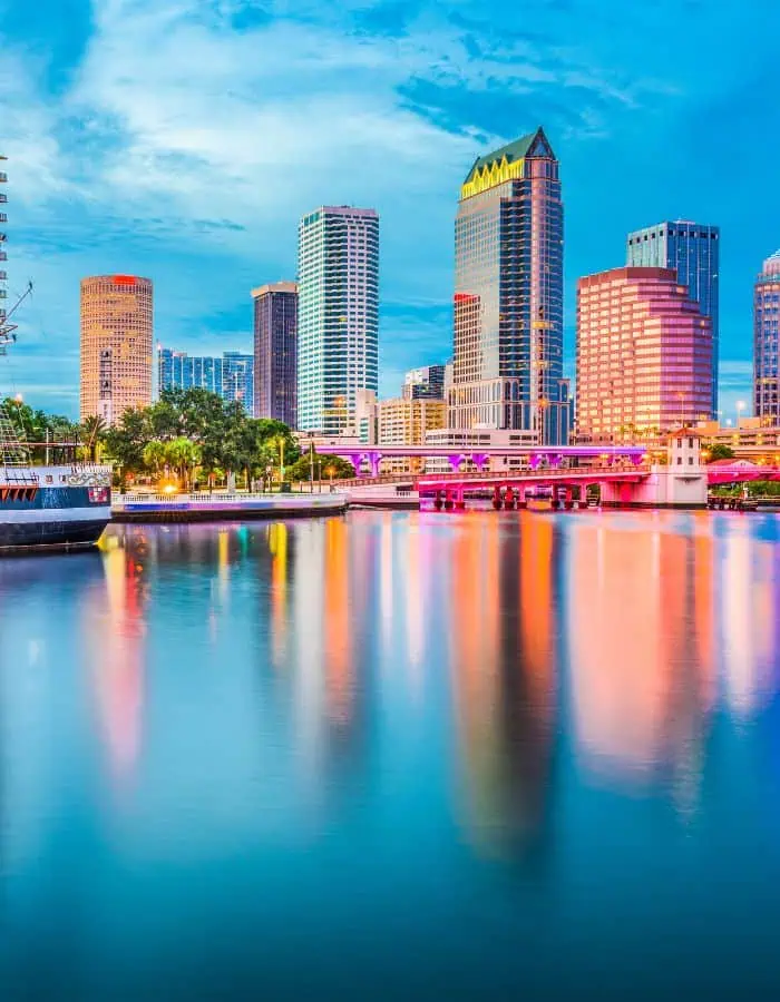 Tampa's skyline with modern high-rise buildings reflects colorfully in the calm river below, as bright pink and orange lights illuminate a bridge across the water. A docked boat rests on the left, completing the lively waterfront scene at dusk.