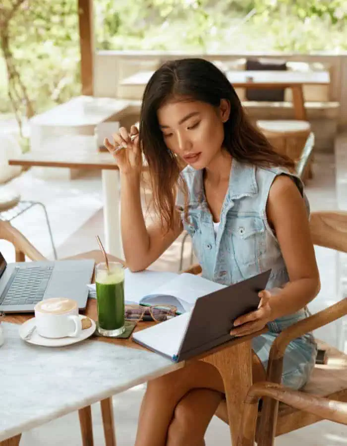 A woman in a sleeveless denim outfit sits at an outdoor café table, focused on a notebook while surrounded by a laptop, a cappuccino, and a green smoothie. The bright, airy setting with natural light and wooden furniture suggests a relaxed yet productive remote work or study environment.