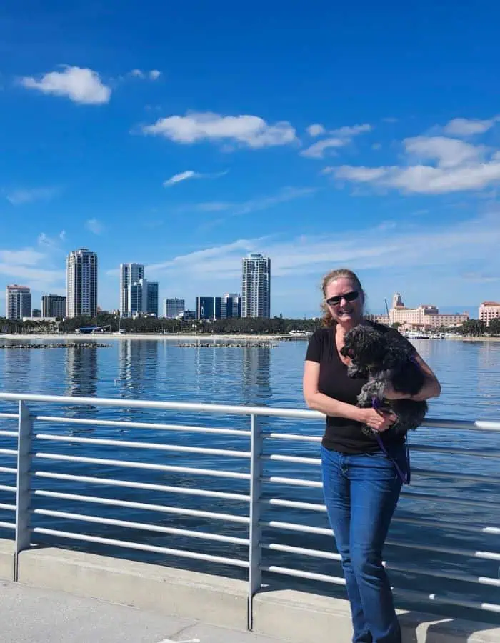 A woman in sunglasses and a black shirt stands on St. Pete Pier holding a small black dog, with a city skyline and high-rise buildings reflected in the calm water behind her. The bright blue sky and scattered clouds enhance the sunny, relaxed atmosphere.