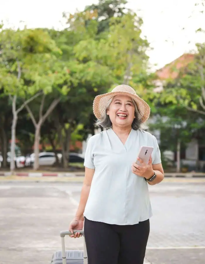 A cheerful older woman wearing a straw hat and light blouse holds a smartphone in one hand and pulls a suitcase with the other. She stands outdoors in a sunny, tree-lined area, radiating excitement and readiness for travel.