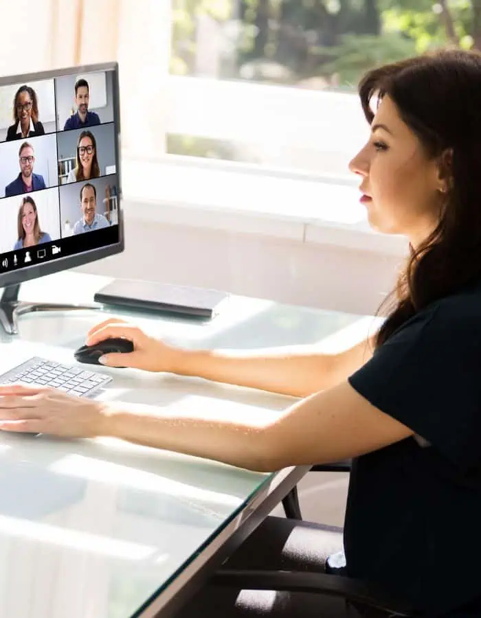 A woman sits at a glass desk in a bright home office, participating in a virtual meeting with seven colleagues shown on her computer screen. Natural light streams through the window, highlighting a modern remote work environment.