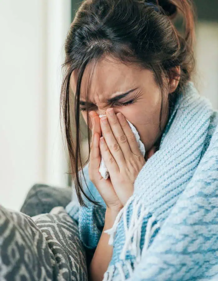 A young woman wrapped in a blue blanket sneezes into a tissue while sitting on a couch, appearing visibly unwell. Her eyes are closed tightly and her posture suggests discomfort, capturing a moment of being sick at home.