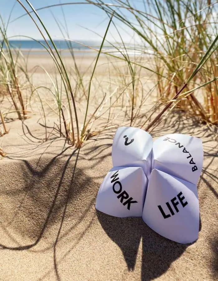 Paper fortune teller with the words “WORK,” “LIFE,” “BALANCE,” and a question mark sits on sandy beach dunes surrounded by tall grass. Ocean and blue sky are visible in the distance, suggesting a focus on work-life balance.