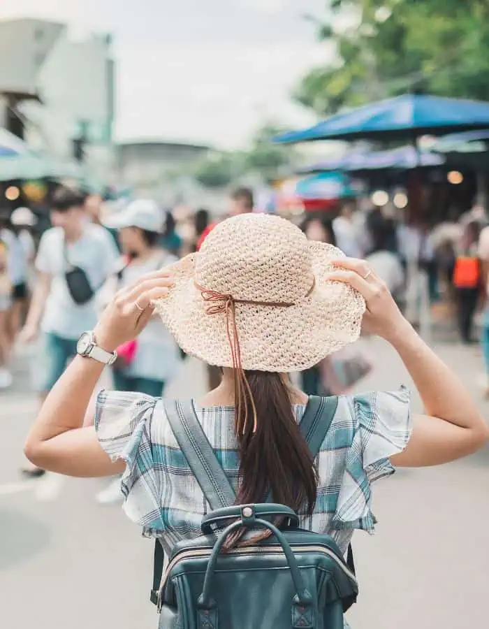 A young woman wearing a straw hat and plaid top is seen from behind as she walks through a bustling outdoor market. She adjusts her hat while carrying a dark backpack, surrounded by a crowd and colorful vendor umbrellas in the background.